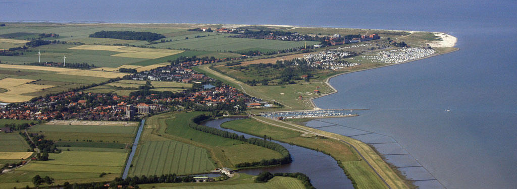Aerial of diked coasline with sea on the right and beach, settlements and green fields on left.