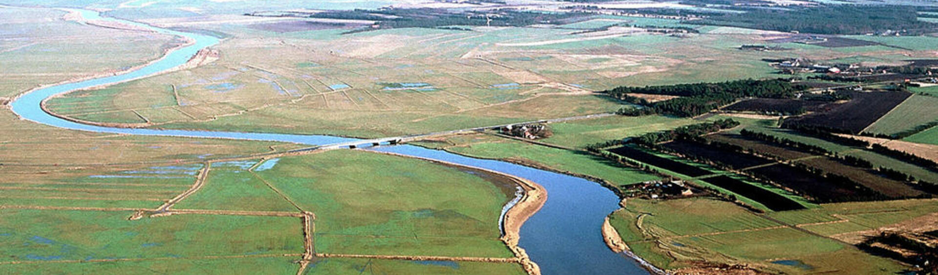 Aerial picture of a winding river with a bridge, green flat areas on the banks and forest and settlement in the distance.