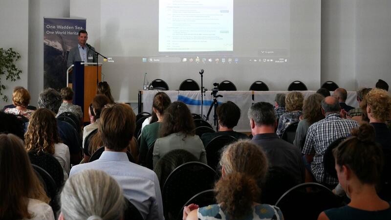 Person standing at a podium desk facing a fully seated conference room. © CWSS.