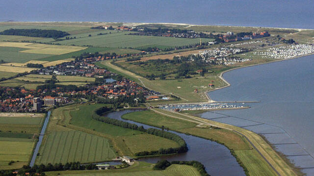 Aerial of diked coasline with sea on the right and beach, settlements and green fields on left.