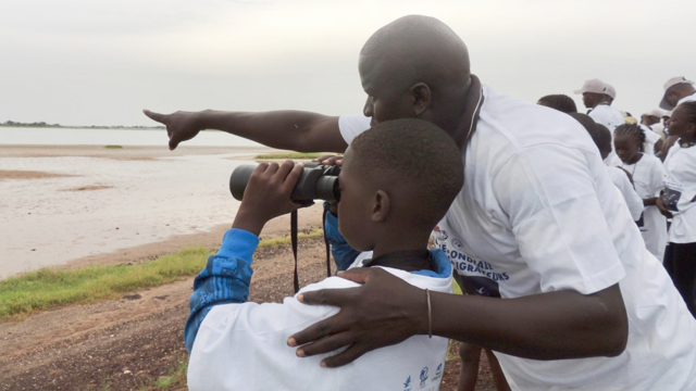 A boy looking through binoculars next to a man pointing at beach and wetlands landscape in distance. © Tim Dodman.
