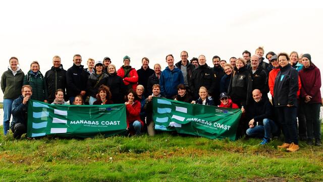 Participants of the MANABAS COAST partner meeting standing and sitting on green grass holding two flags with the MANABAS COAST project logo. © Ricardo Fijma.