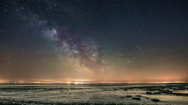 Dark starry sky above exposed mudflats with lights of coast in the distance. © Raymond Kamstra.