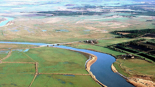 Aerial picture of a winding river with a bridge, green flat areas on the banks and forest and settlement in the distance.